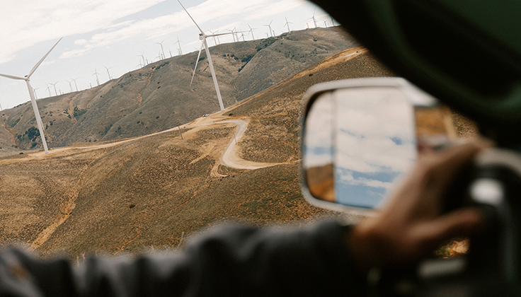 A view from the inside of a vehicle shows wind turbines along a dry hillside. 