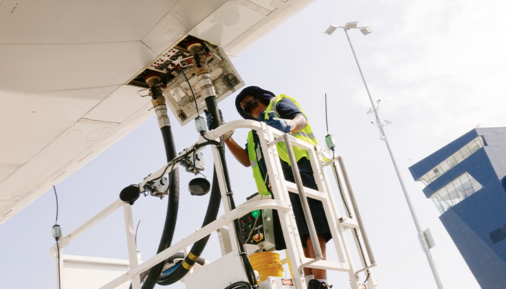 A person wearing safety gear is shown operating machinery that provides fuel to a large aircraft. 