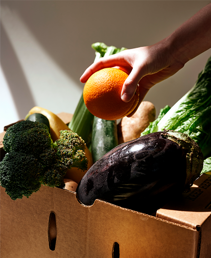 A hand pulling an orange out of a paper crate filled with fresh produce.