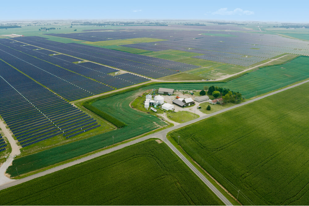 Large, green fields with solar panels and building structures in the middle are shown from above. 