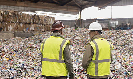 Two people stand in front of recyclable materials in a recycling facility.